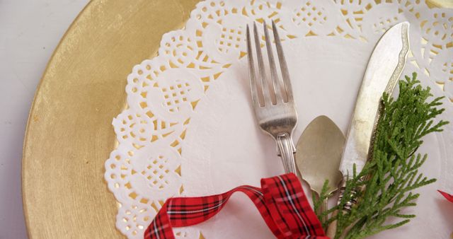 A festive table setting features a gold charger plate and elegant cutlery, with copy space. The red ribbon and greenery add a touch of holiday cheer to the dining presentation.