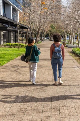 College friends walking tree-lined campus path carrying skateboard and backpack