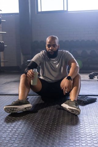 African man sitting on gym floor holding shaker bottle during post-workout recovery