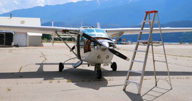 Small airplane with propeller parked on tarmac next to a ladder at airport. Ideal for illustrating topics related to aviation, small aircraft maintenance, airport activities, pilot training, and aviation industry behind-the-scenes.