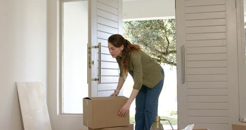 Woman moving into new home lifting boxes in sunlit entryway near open double doors