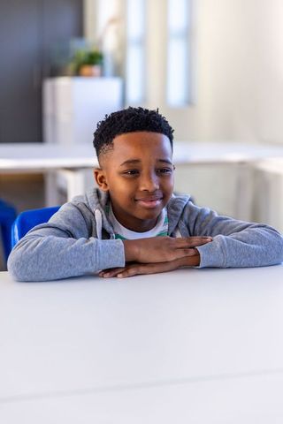 Thoughtful african american schoolboy resting at classroom desk wearing hoodie and smiling