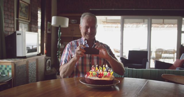 Caucasian senior man taking photo of birthday cake using smartphone at home. Retirement, senior lifestyle, happiness, domestic life, birthday, celebration, communication and wine making, unaltered.