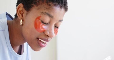 Young woman wearing orange hydrogel under-eye patches smiling with eyes closed at vanity