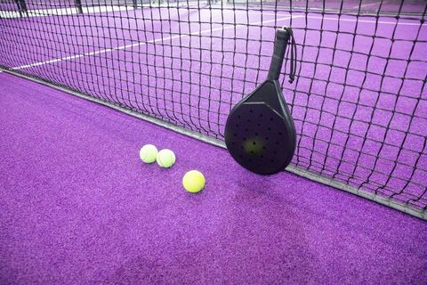 Black padel racket leaning on net with three yellow balls on vibrant purple court