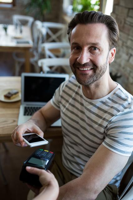 Man sitting in a coffee shop, smiling while making a contactless payment using his mobile phone. Ideal for illustrating modern payment methods, technology in everyday life, and customer satisfaction in a casual setting. Can be used for articles on digital transactions, mobile payment apps, and the integration of technology in daily activities.