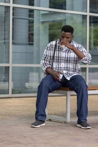 African man sitting on bench sipping coffee checking smartphone outside urban glass facade