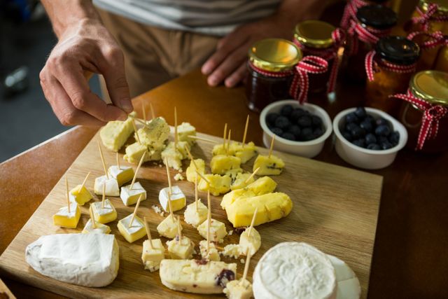 This image shows a close-up of a staff member arranging various types of cheese on a wooden board in a grocery shop. Ideal for use in marketing materials for gourmet food shops, delicatessens, or dairy product promotions. It can also be used in blogs or articles about cheese varieties, food preparation, or retail display techniques.