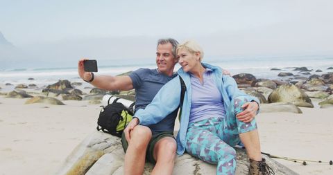 senior couple sitting on a rock near the beach taking a selfie. visible are their backpacks and hiking poles, depicting an adventurous and active lifestyle. perfect for promoting senior fitness, outdoor activities, adventure travel, and the joy of exploration together.