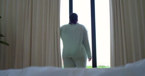Middle-aged african-american woman drawing curtains in pajamas by bedroom window