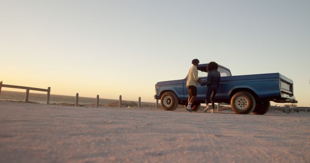 Couple leaning on a vintage blue pickup truck with a sunset backdrop at the beach. Used for themes of romance, nostalgia, travel mood, outdoor adventure, or relaxation. Ideal for marketing travel agencies, lifestyle blogs, and advertisements portraying romantic escapes or road trips.