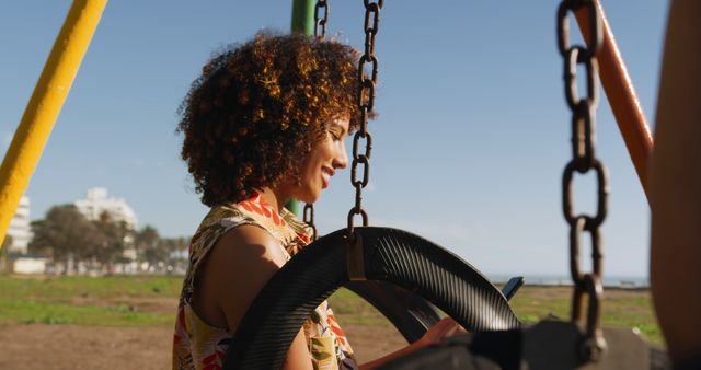 Happy biracial woman using smartphone sitting on swing at sunny playground, copy space. Communication, summer, vacations, lifestyle and free time, unaltered.