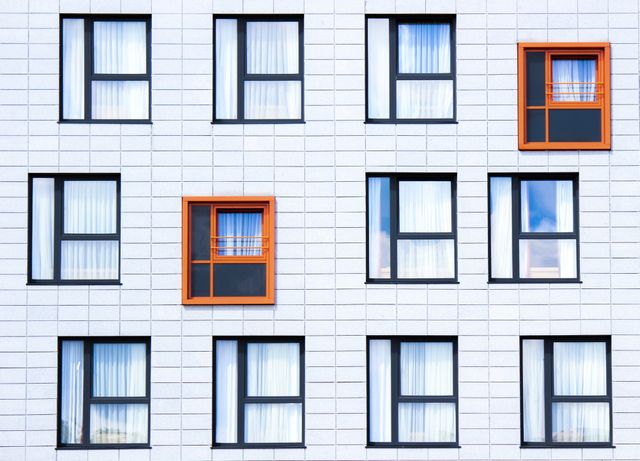 Shows a white modern building facade with multiple black-framed windows. Features unique orange window accents among the uniform reflections. Ideal for illustrating urban life, modern architecture, design concepts, and contemporary real estate developments.