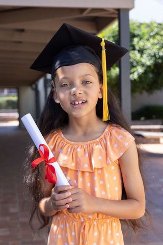 Young girl holding diploma wearing mortarboard with yellow tassel in school corridor