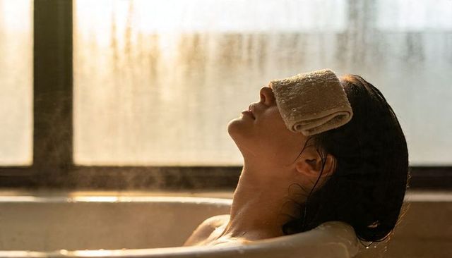 Sunlit bathtub relaxation: woman reclining with warm towel over eyes, steam glow