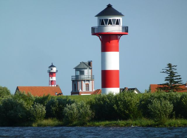 Lighthouses with red and white stripes standing on the riverside surrounded by greenery. Scenic backdrop with clear blue sky and natural elements offers maritime charm. Ideal for uses related to coastal living, travel, navigation, and nature-themed projects.