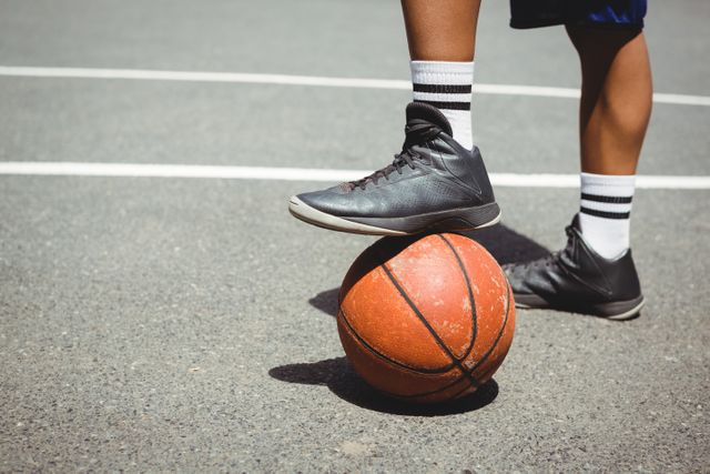 This image shows an athlete standing on an outdoor basketball court with one foot resting on a basketball. The focus is on the lower body, highlighting the athletic shoes and socks. Ideal for use in sports-related content, fitness blogs, training programs, and advertisements for athletic footwear or active lifestyle promotions.