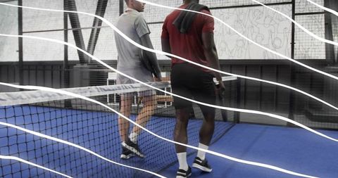 Two teammates walking across indoor padel court carrying paddles passing net on blue court