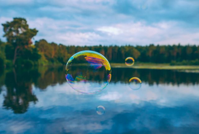 Image features vibrant soap bubbles floating gently above a calm lake with a serene forest and cloudy sky in the backdrop. Could be used to illustrate themes of peace, nature, childhood, relaxation, or delicate beauty.