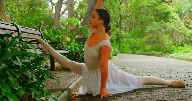 Woman demonstrating flexibility and fitness as she stretches in a scenic park with lush greenery and sunlight filtering through trees. Useful for themes of health, fitness, ballet, relaxation, outdoor activities, and performance arts promotions.