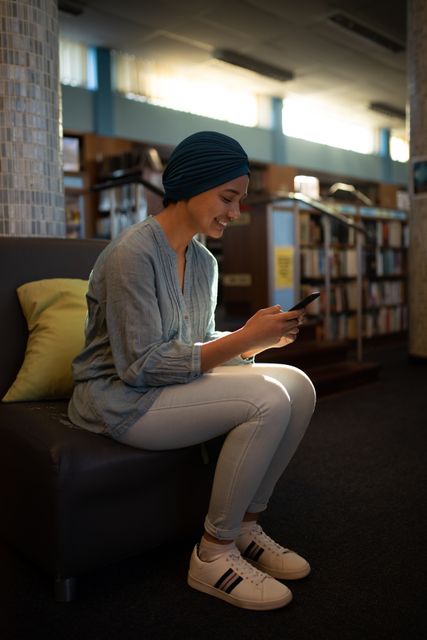 Young biracial female student wearing a dark blue hijab is sitting on a seat in a library, text messaging on her phone. She is dressed casually in a light blue top and white pants, with a pillow beside her. The library shelves filled with books are visible in the background. This image can be used for educational content, promoting diversity and inclusion, technology in education, or student life themes.