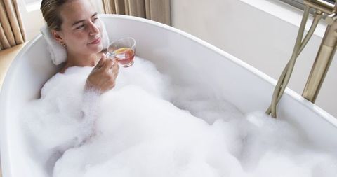 Woman relaxing in bubble bath holding cup of lemon tea in minimalist neutral bathroom