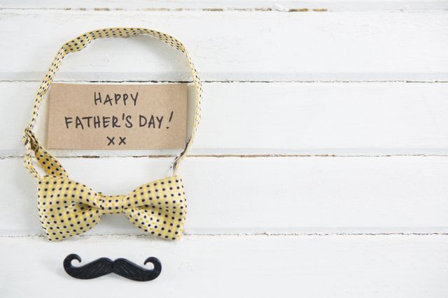 Perfect for Father's Day promotions, greeting cards, and social media posts. The image features a handwritten 'Happy Father's Day' message on a card, accompanied by a yellow polka dot bow tie and a black mustache on a white wooden background. Ideal for conveying love and appreciation for fathers.