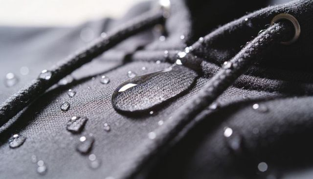 Macro water beading on dark hoodie fabric near drawstring and grommet showing hydrophobic finish