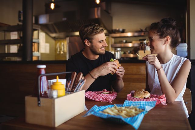 Young couple smiling and interacting while enjoying burgers and fries in a cozy bar. Ideal for use in advertisements for restaurants, fast food promotions, or lifestyle blogs focusing on dining out and social interactions.
