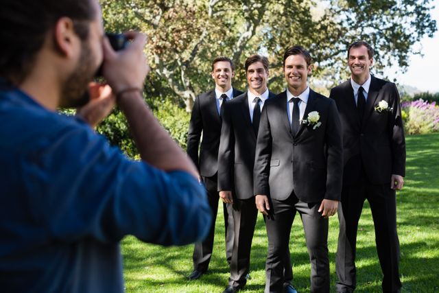Groom and groomsmen posing for a photographer in a park, all dressed in formal black suits with ties. They are smiling and standing in a line, with greenery and trees in the background. Ideal for use in wedding planning materials, photography portfolios, or articles about wedding traditions and celebrations.