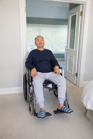 Senior chinese man sitting in wheelchair at bedroom doorway wearing slippers and sweatpants in dayli