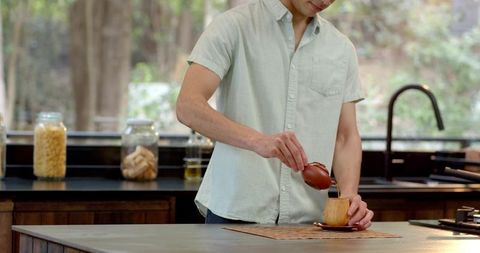 Man pouring tea from clay teapot into wooden cup in modern kitchen