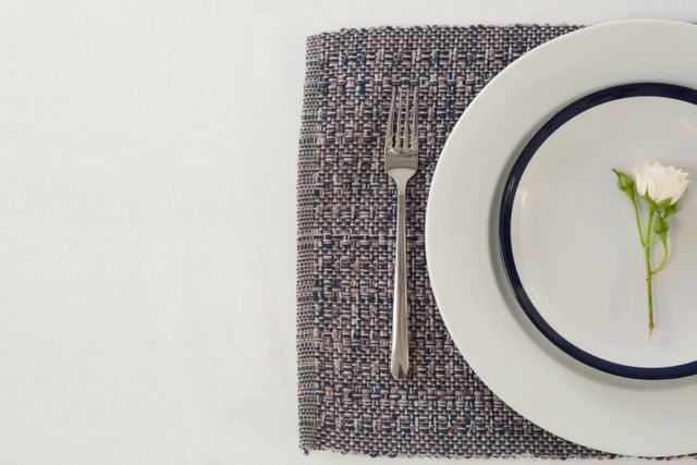 Overhead view of an elegant table setting featuring a white plate with a small white rose, a fork placed on a woven placemat. Ideal for use in articles or advertisements about fine dining, home decor, hospitality, or minimalistic table settings.
