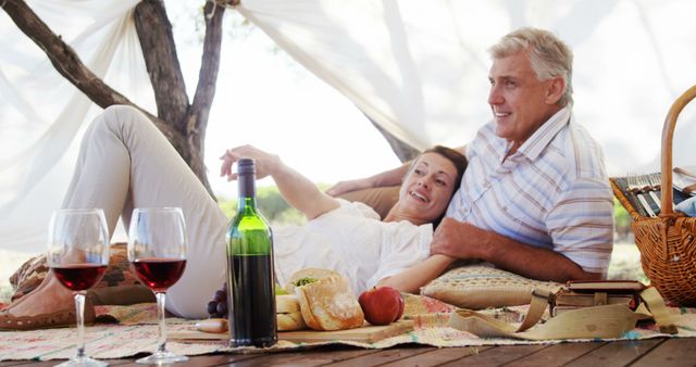 Senior couple reclining on picnic blanket outdoors with wine, bread, wine glasses, and food basket around. Great for retirement, leisure, wholesome living, and outdoors lifestyle themes.
