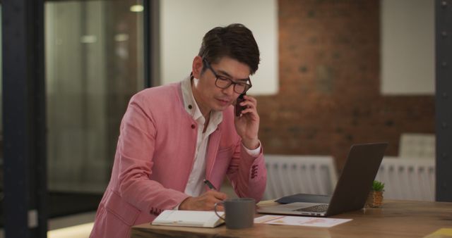 A young Korean businessman wearing a pink blazer is on a phone call while taking notes and working on a laptop in a modern office setting. This image is useful for business, communication, technology, or professional lifestyle themes in marketing materials, presentations, or websites.