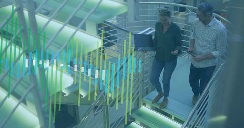 Two colleagues walking and collaborating on modern staircase with laptop and smartphone