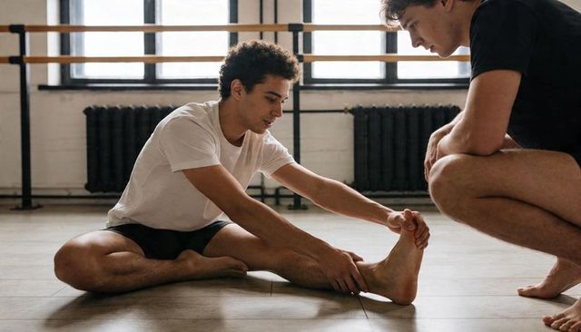 Dancer stretching hamstring with coach guiding in sunlit ballet studio for flexibility training