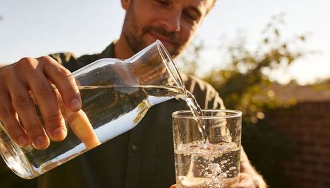 Man pouring refreshing water from glass carafe into tall glass in sunlit backyard