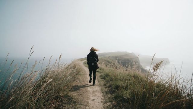 Woman walking coastal cliff path through misty windblown landscape solo hike