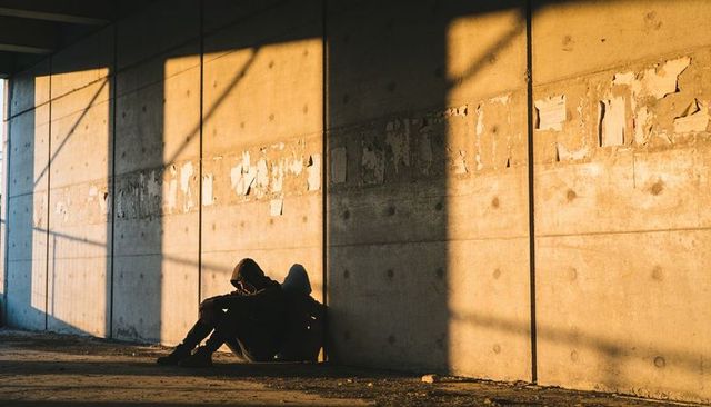 Two hooded adults sitting against sunlit concrete underpass wall casting grid shadows