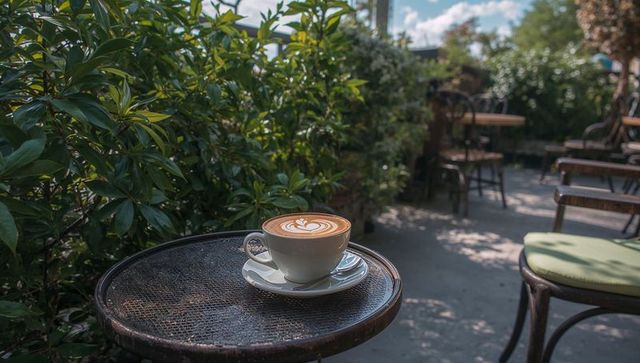Sunlit Latte Sitting on Metal Bistro Table in Green Outdoor Café Patio