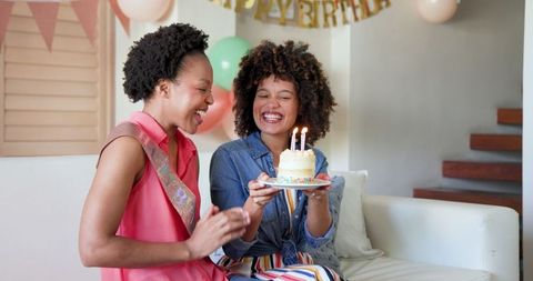 Two joyful women are smiling and holding a birthday cake with burning candles in a cheerful atmosphere at home. A fun banner and balloons enhance the festive environment, conveying warmth and friendship. Ideal for promotion of birthday parties, celebrating diverse friendships, or social gathering events.