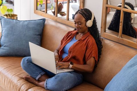 African american woman working remotely on laptop with headphones on cozy living room sofa