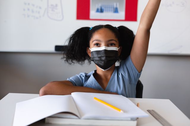 Biracial elementary schoolgirl wearing mask while sitting with hand raised at desk in classroom. unaltered, education, intelligence, protection, safety, coronavirus, disease and school concept.