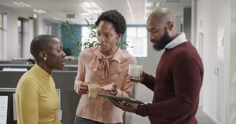 three colleagues are gathered in an office, engaging in a productive conversation while using a tablet. they hold coffee mugs, symbolizing a casual yet interactive working environment. this image is excellent for content related to teamwork dynamics, productive meetings, or office management materials.