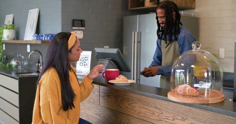 Indian woman in mustard sweater ordering coffee at modern cafe counter with croissant
