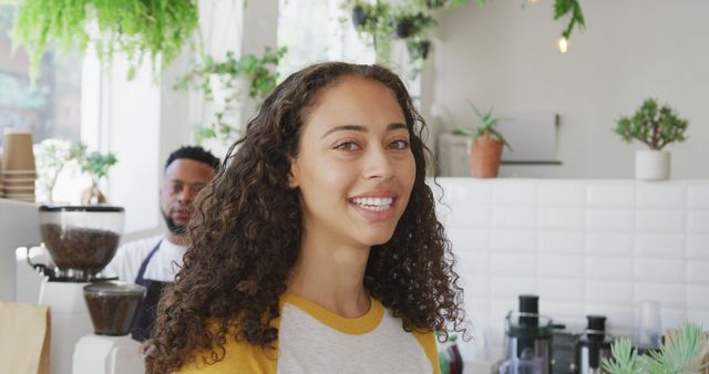 Female cafe worker smiling, standing in front of counter with plants hanging and placed around. Ideal for promoting cafes, hospitality training materials, advertisements focusing on customer service, or illustrating warm, friendly business environments.