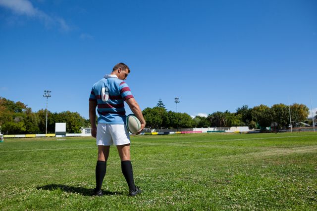 Rugby player standing on a green field, holding a ball, with a clear blue sky in the background. Ideal for use in sports-related content, advertisements for athletic gear, fitness and training programs, or promoting outdoor activities and team sports.