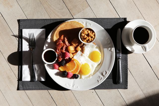 A breakfast plate with sunny-side up eggs, toast, bacon, sautéed potatoes, pancakes, fruit including berries and orange slices, accompanied by black coffee on a wooden table. Ideal for illustrating healthy and complete breakfast meals, weight management programs, restaurant menus, or morning advertisements.