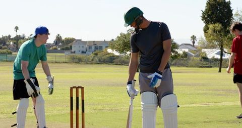 Cricket batsman practicing on sunny park pitch with helmet, pads and wooden stumps